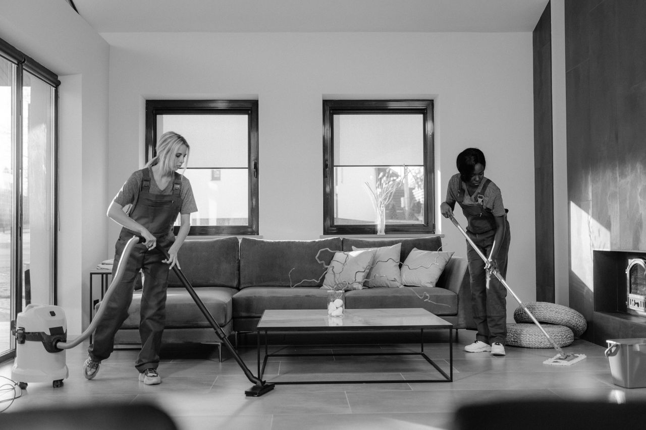 services-09 Two women in work clothes cleaning a modern living room using a vacuum and mop.