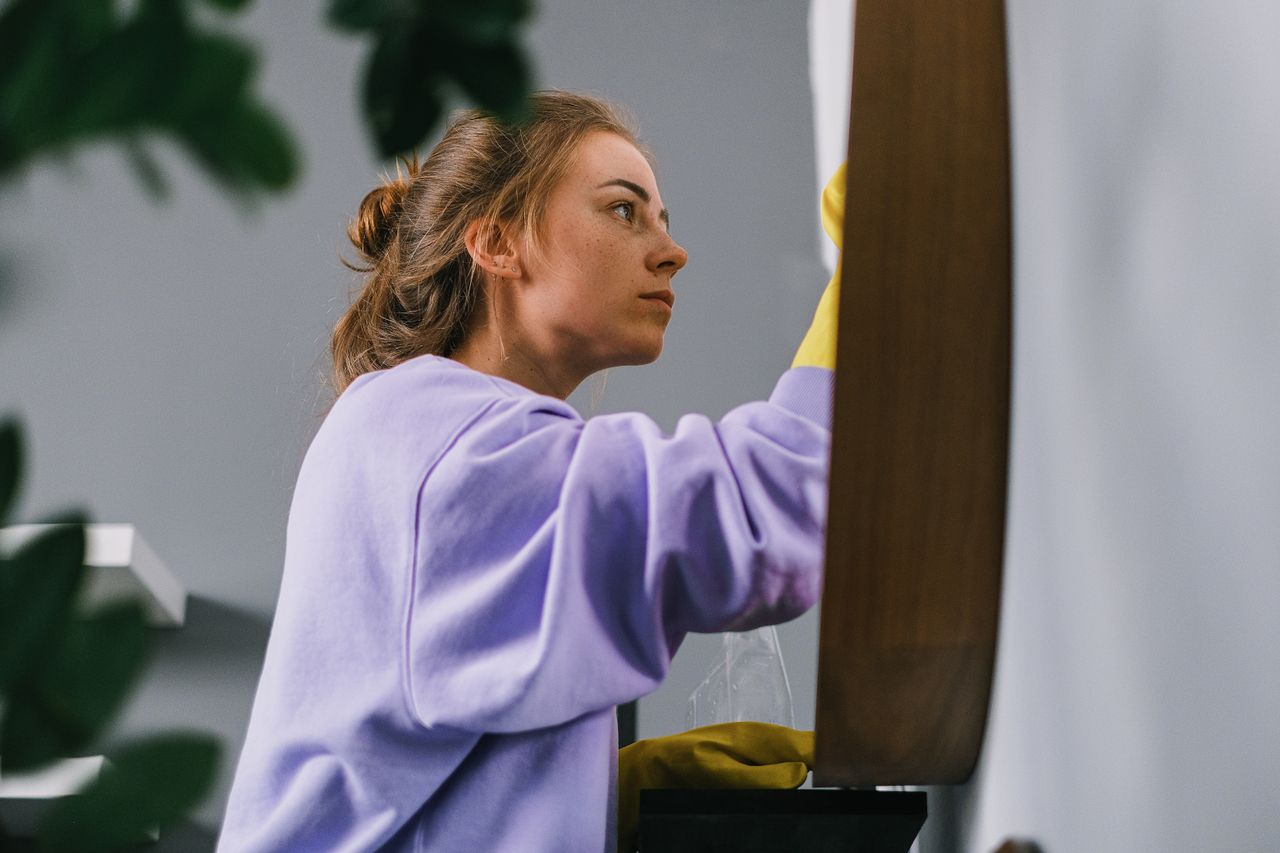 hero-02 Side view of young focused female in latex gloves standing and cleaning mirror in light room in daytime
